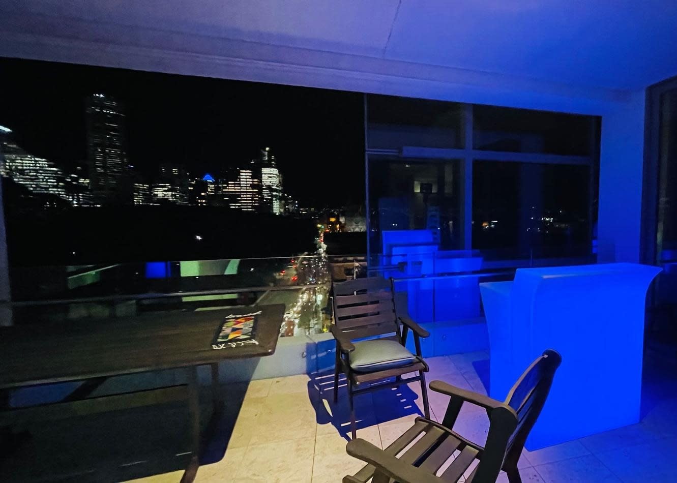 A balcony with a straight bar hire and a view of the city, illuminated by blue lights.