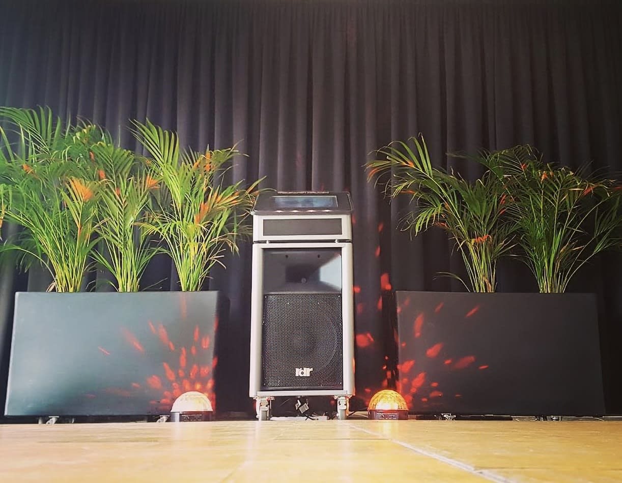 A stage with a speaker and plants in front of it, featuring Pkg 2: Jukebox & Karaoke Hire.