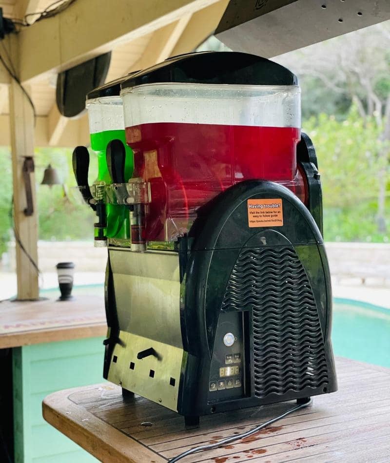 Description: A soda machine sitting on a table next to a pool, offering Slushie Machine Hire with a package that includes *240 drinks*.