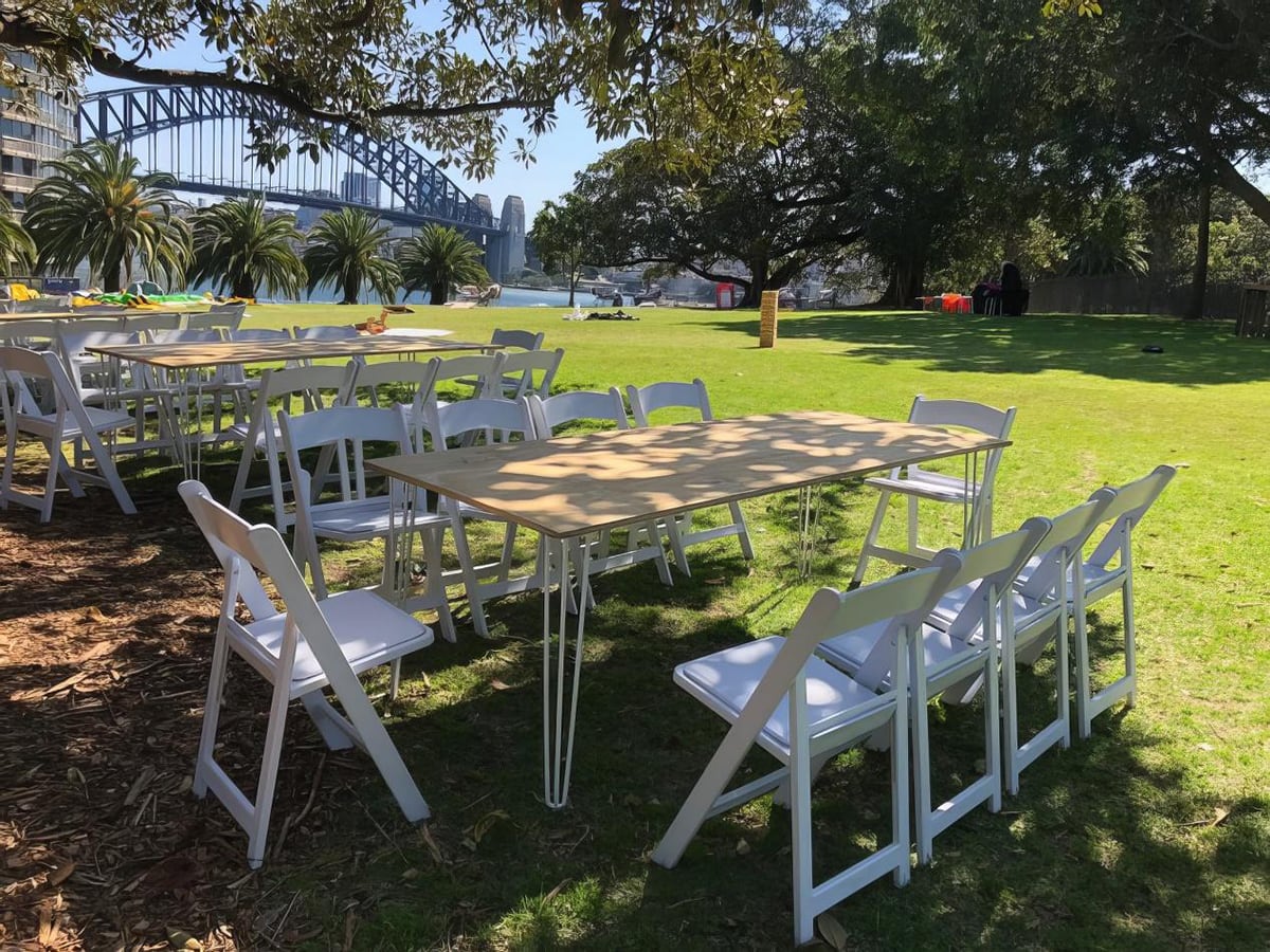 Our timber top hairpin table with some white stackable chairs being used at an outdoor park next to the harbour bridge