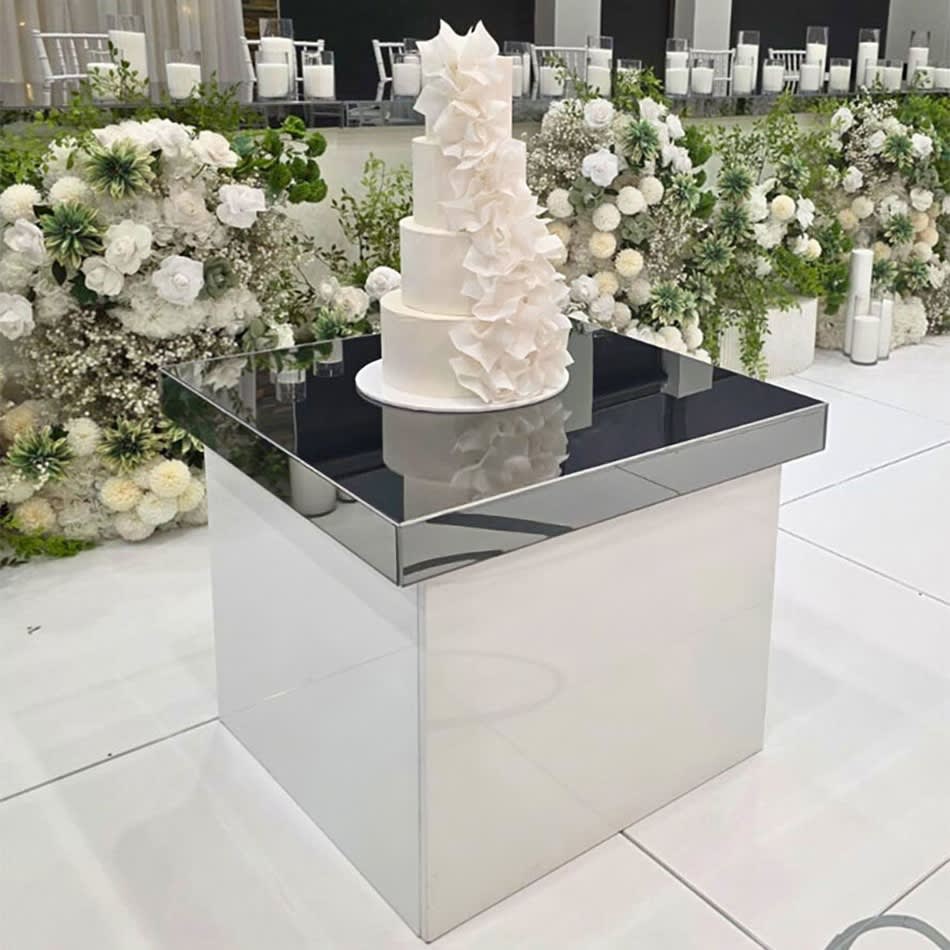 Three-tier white wedding cake with cascading sugar flowers displayed on a reflective gloss cake table, styled with white floral arrangements and modern décor.