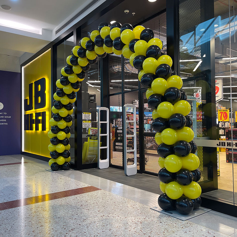 Black and yellow balloon arch framing the entrance of a JB Hi-Fi store in a shopping centre, styled for a retail promotion or grand opening.