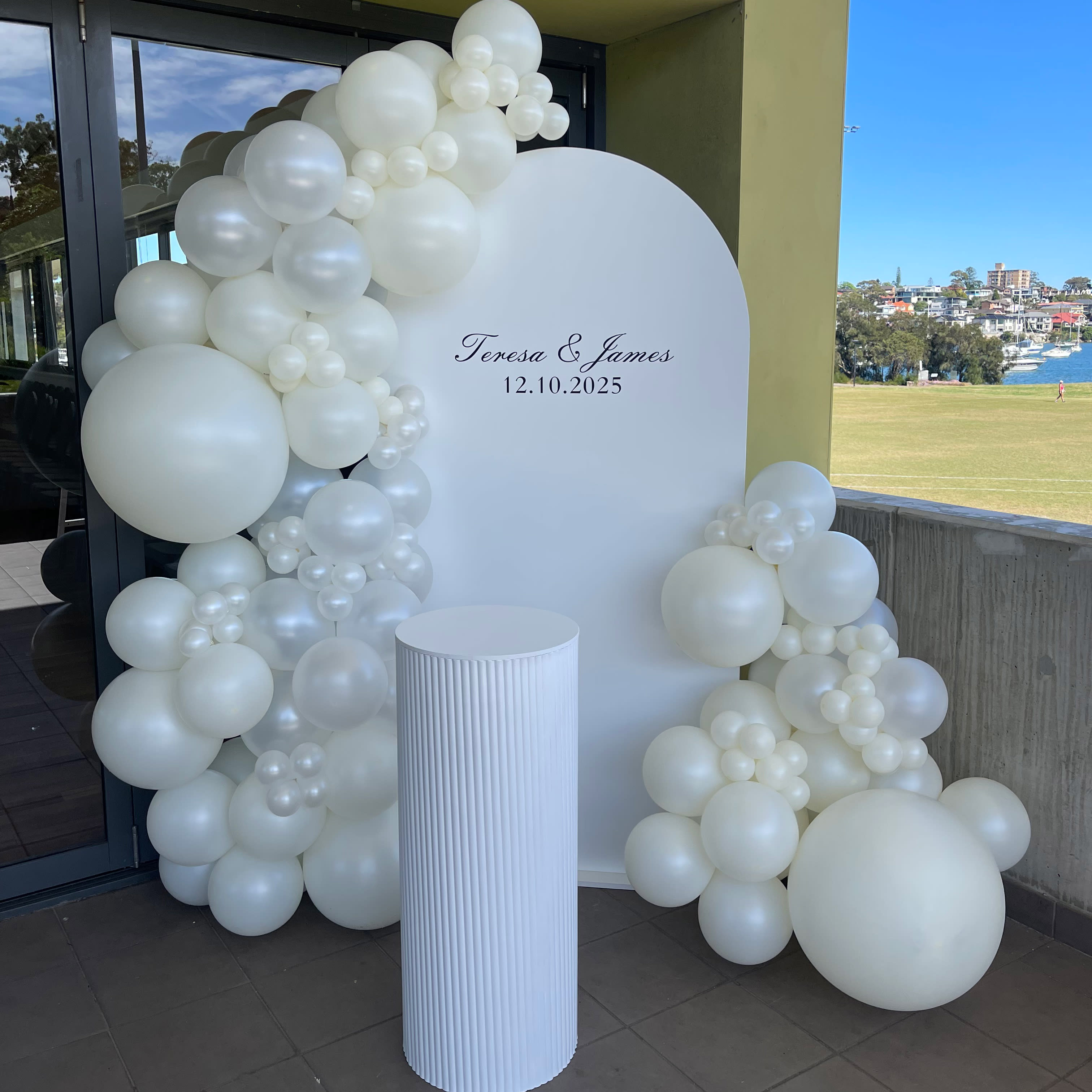 White balloon garland styled around a white round backdrop with personalised lettering, paired with a matching white plinth for a clean and elegant event display.