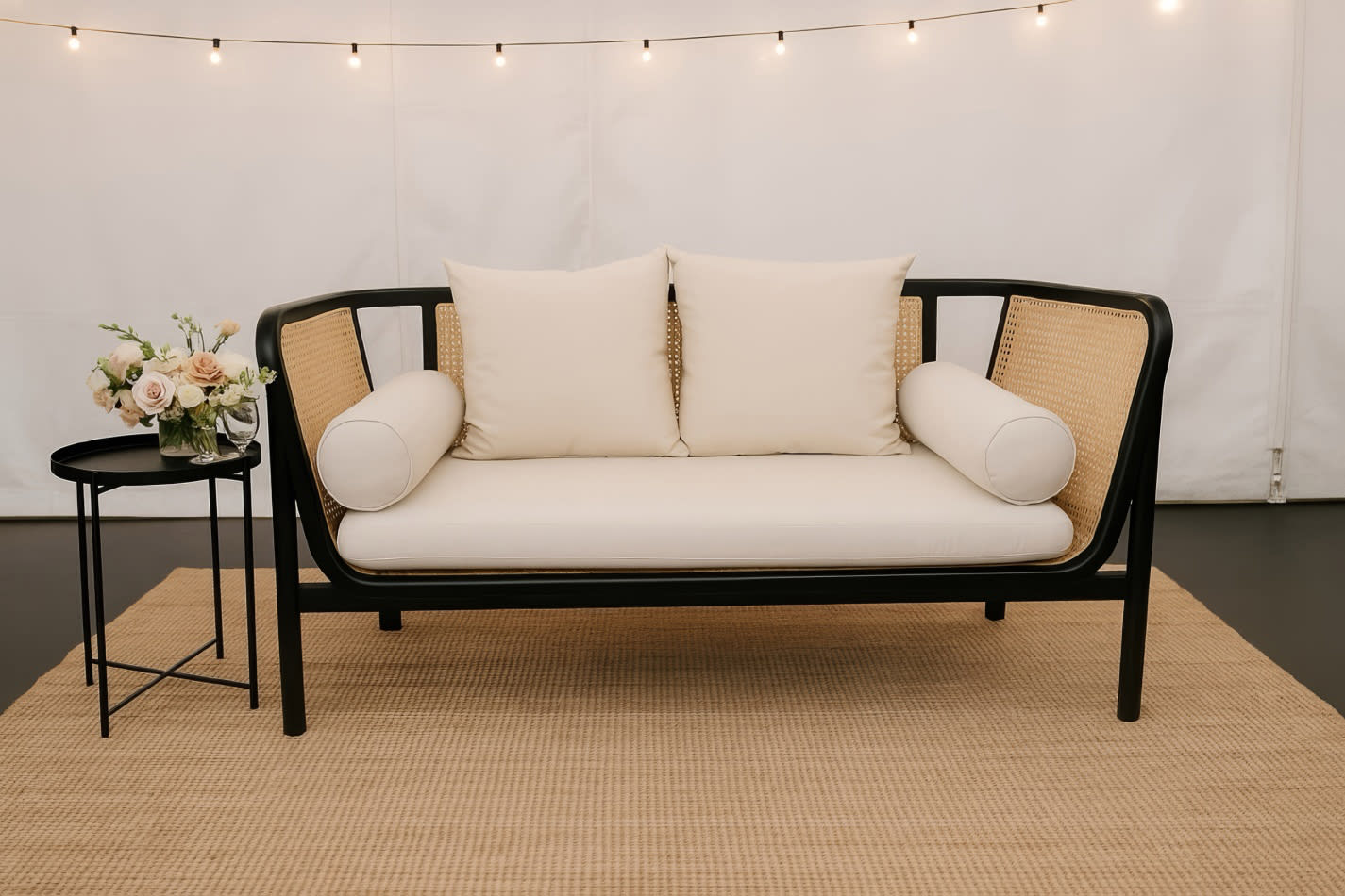 Black round side table styled beside a rattan lounge with cream cushions, set on a woven rug with soft decorative lighting for an elegant event seating setup.