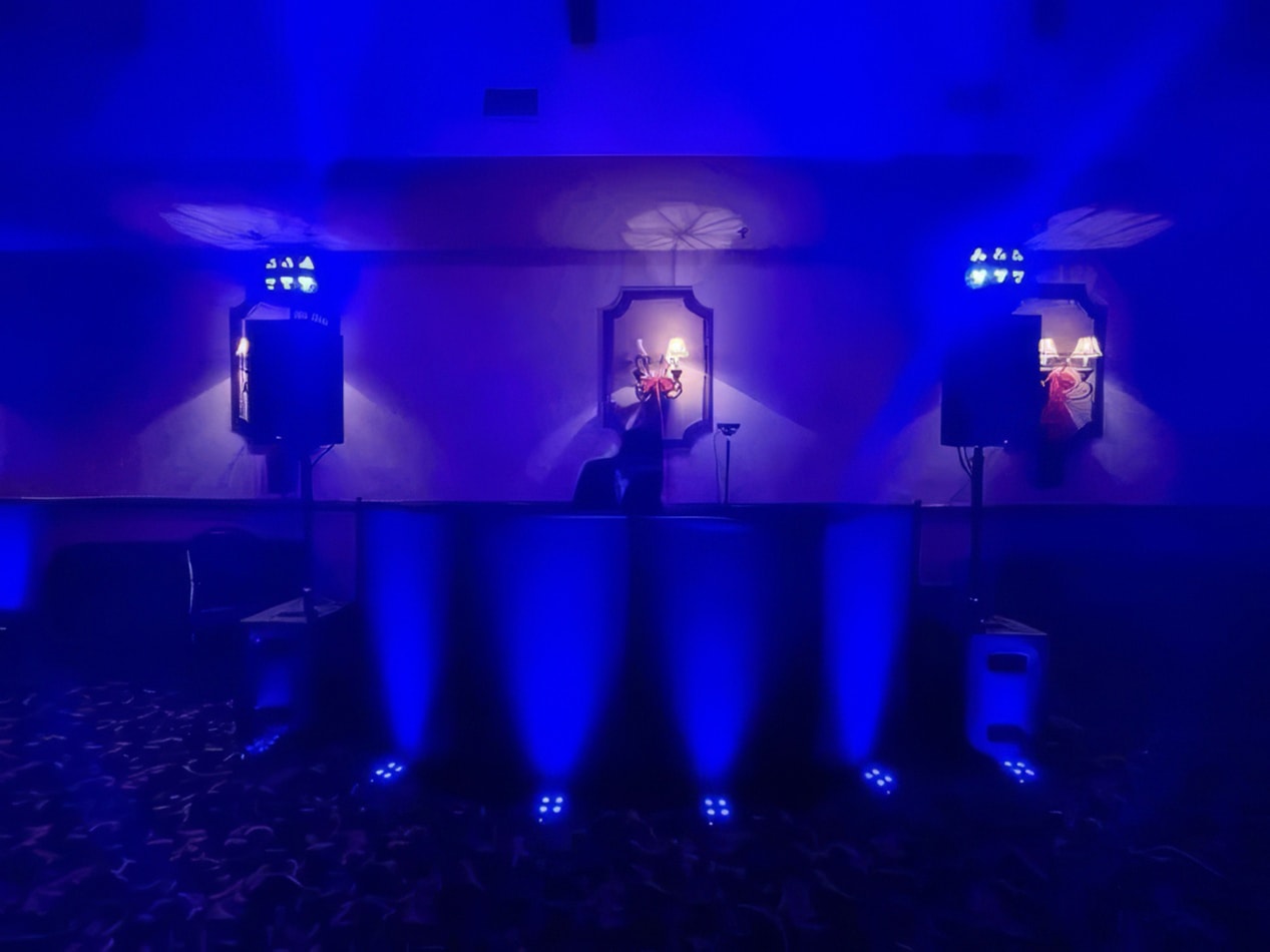 Blue battery-operated par can uplights illuminating a DJ booth and speakers in a dark event space, creating a vibrant atmospheric wash across the room.