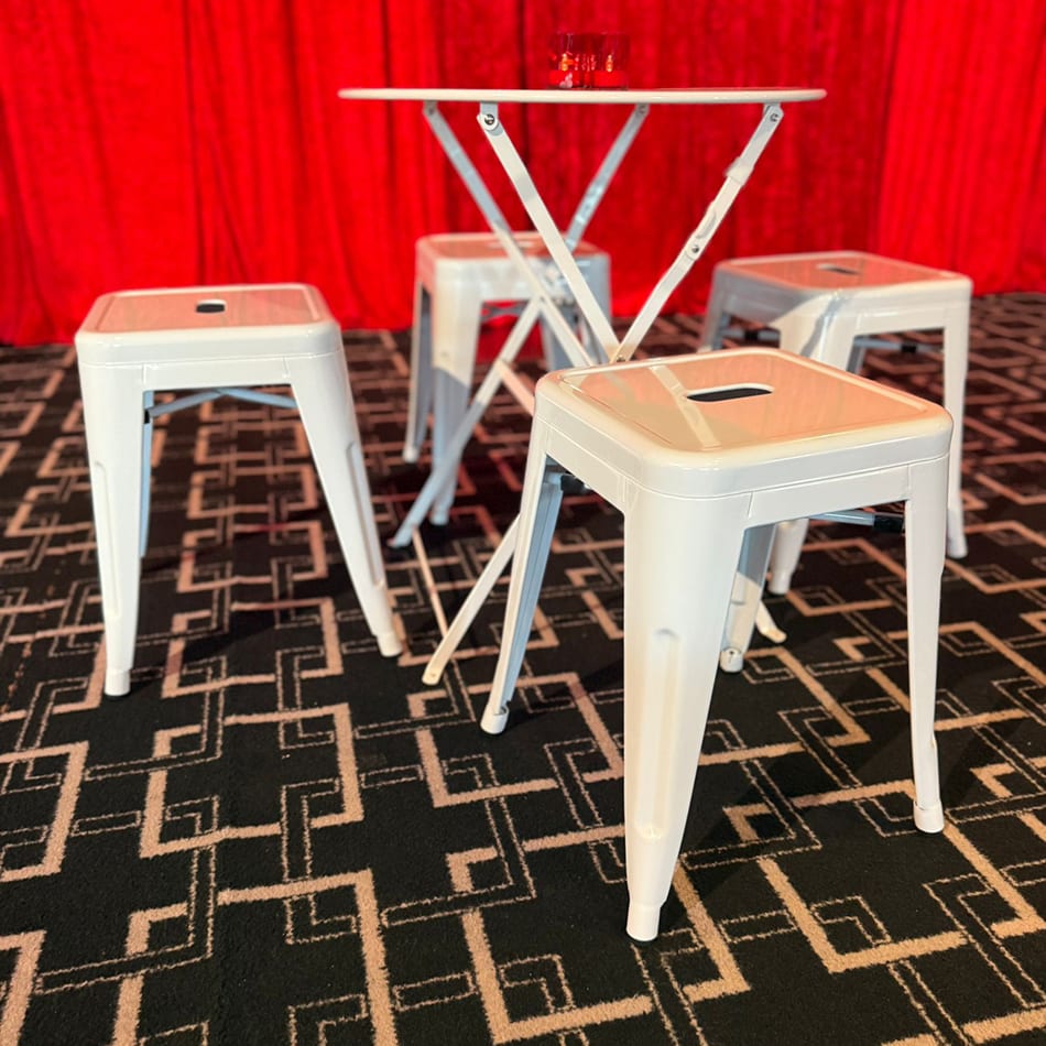White metal bar table with four matching stools arranged on a geometric black carpet in front of a red curtain backdrop at an event venue.