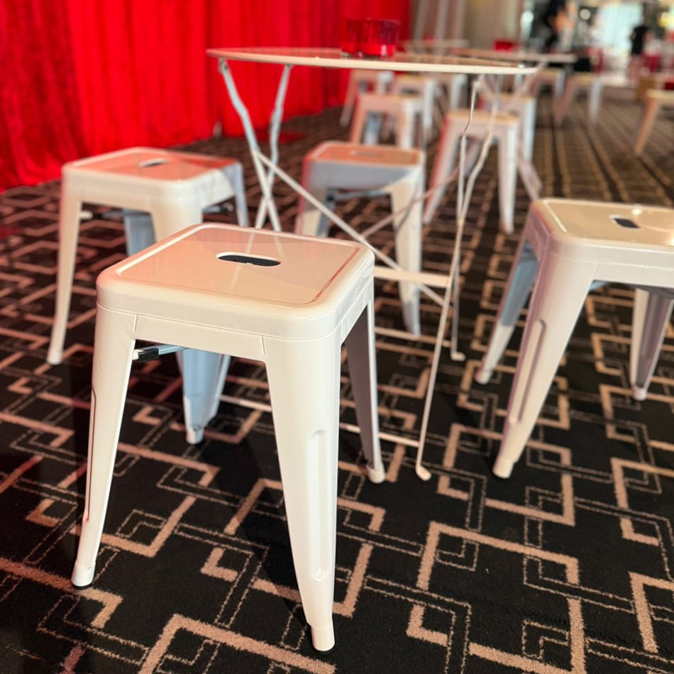 Close-up of white metal bar stools and matching table set on patterned carpet with a red curtain backdrop at an indoor event.