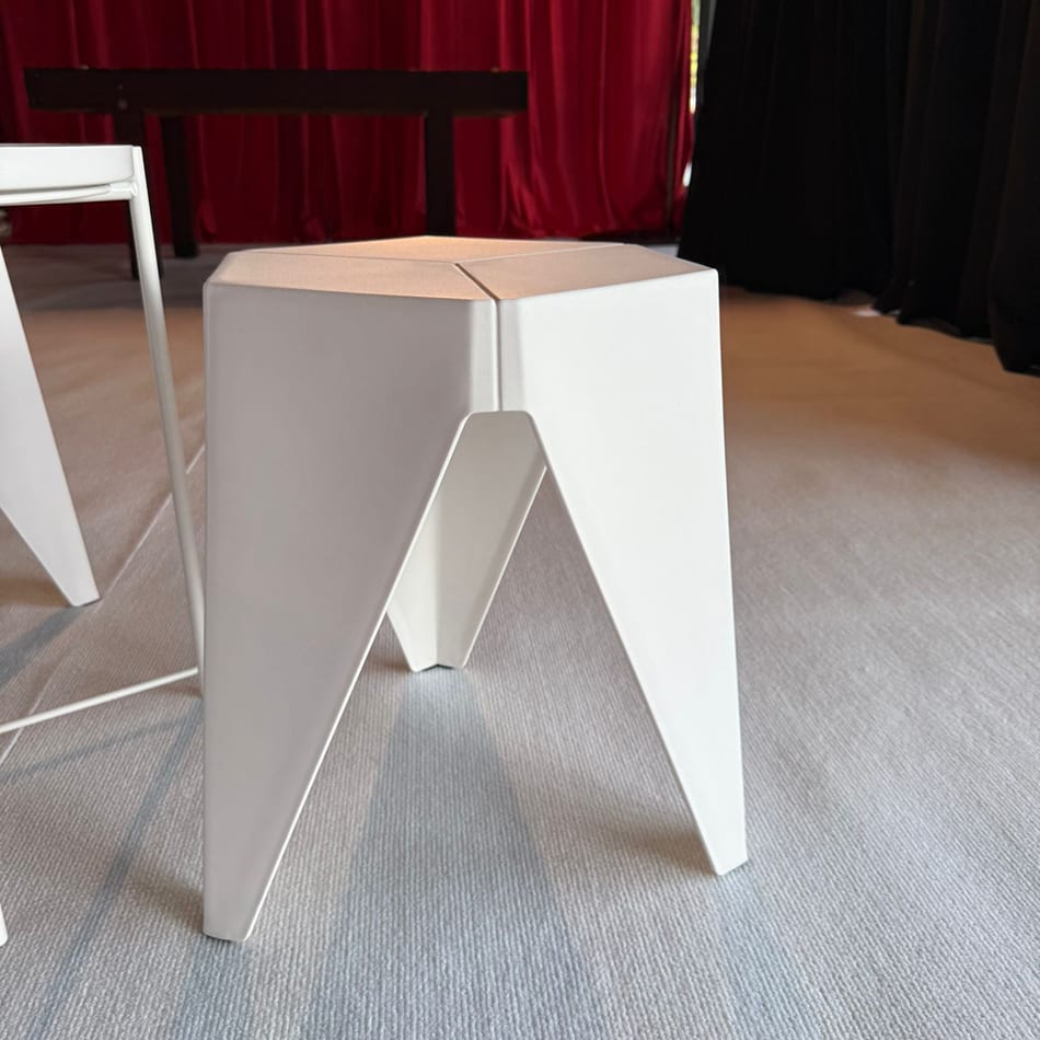 White Puzzle Stool with geometric design positioned on a light grey carpet in front of a red curtain at an indoor event venue.