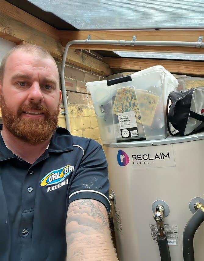 Plumber standing beside a newly installed heat pump hot water storage tank with connected pipework and fittings inside a service area