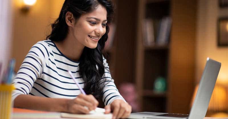 woman writing in notebook while using laptop