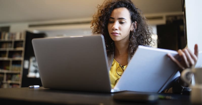 woman at desk with computer looking at notebook