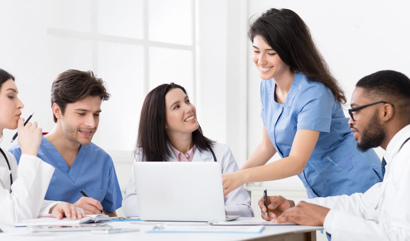 Five nurses stand outside a windowed building and smile at the camera.