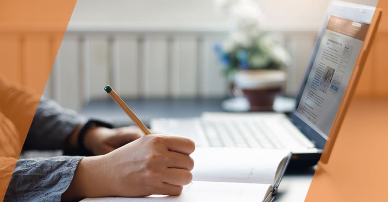 person's hand writing in a book by a laptop on their desk