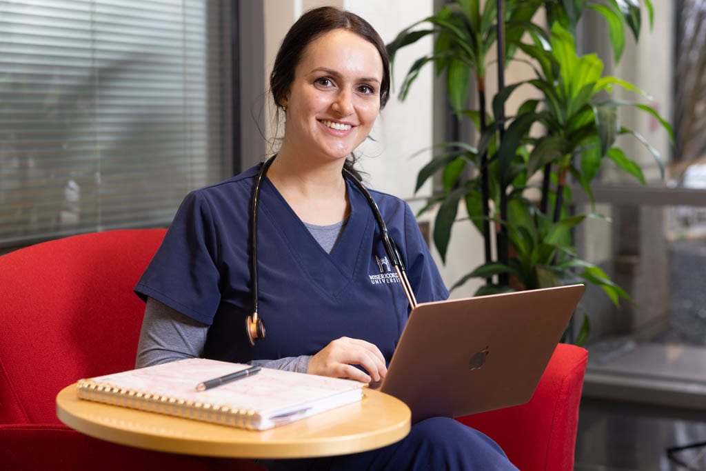 nursing student in lounge with laptop and books smiling at camera