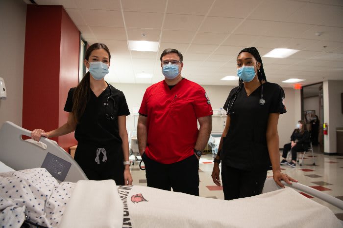 3 nursing students in masks standing over clinical bed looking into the camera