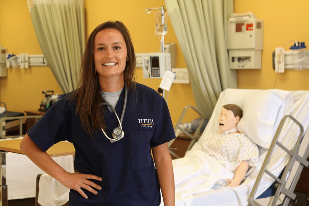 Utica student smiling in front of manikin in hospital bed