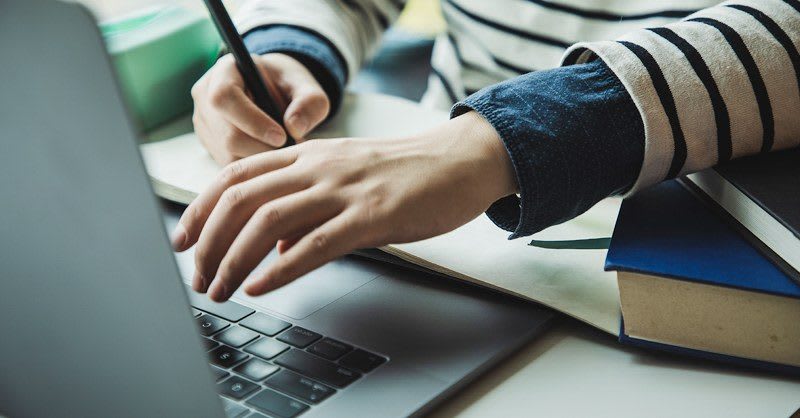 Hands typing on computer and taking notes with pen