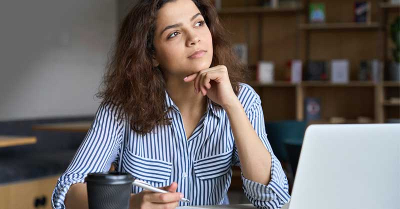 Woman sitting at desk thinking, while working on laptop