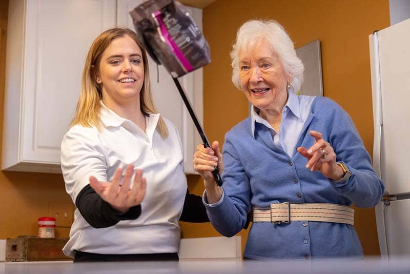 OT student helping elderly woman lift bag in kitchen