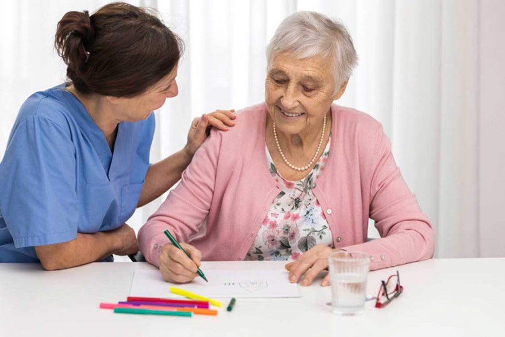 woman in scrubs sitting with elderly woman at table drawing on paper