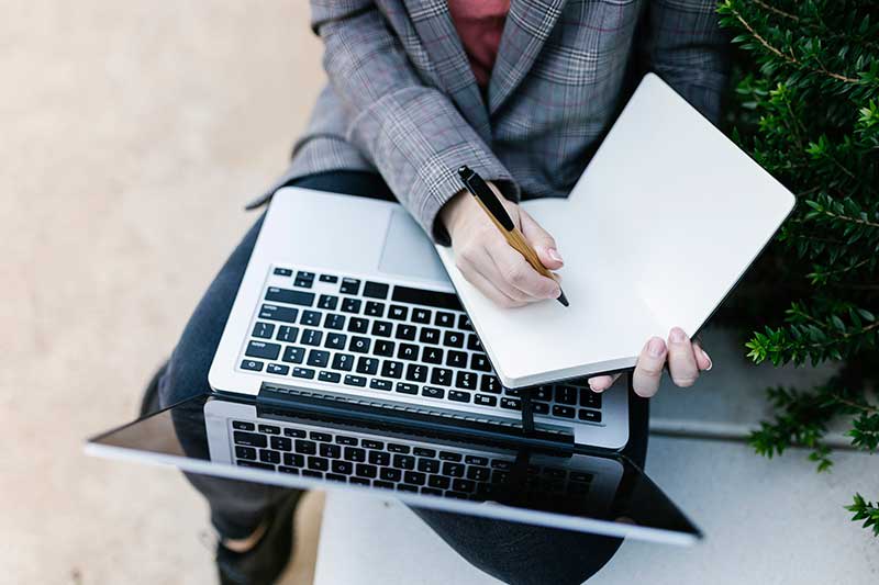 person sitting down with a laptop and writing in journal