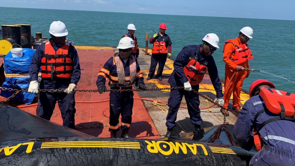 A team of oil spill response professionals wearing life jackets and helmets perform a containment drill on a vessel in Suriname. The exercise demonstrates best practices in marine pollution control and preparedness.