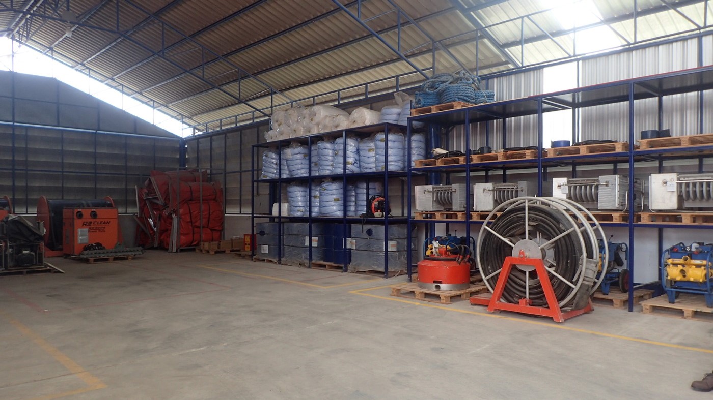 A well-organised storage facility housing oil spill response equipment, including booms, hoses, and skimmers, all systematically stored on blue metal shelves.