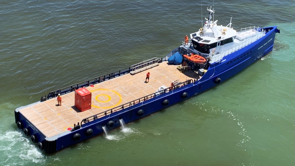 A blue oil spill response vessel with crew members on deck, equipped with emergency equipment and a helipad, positioned for rapid deployment in Suriname’s waters.