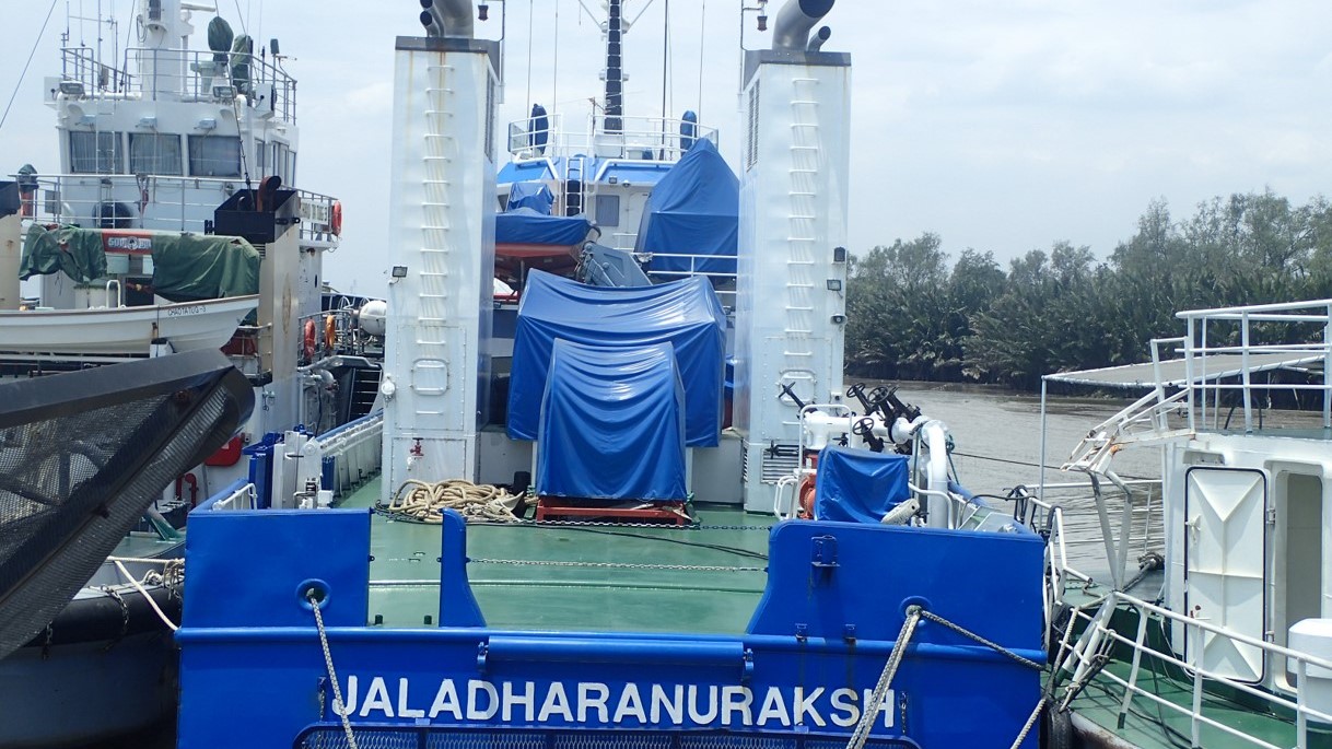 A blue oil spill response vessel docked at a riverbank, equipped with containment and recovery equipment covered with protective tarps.