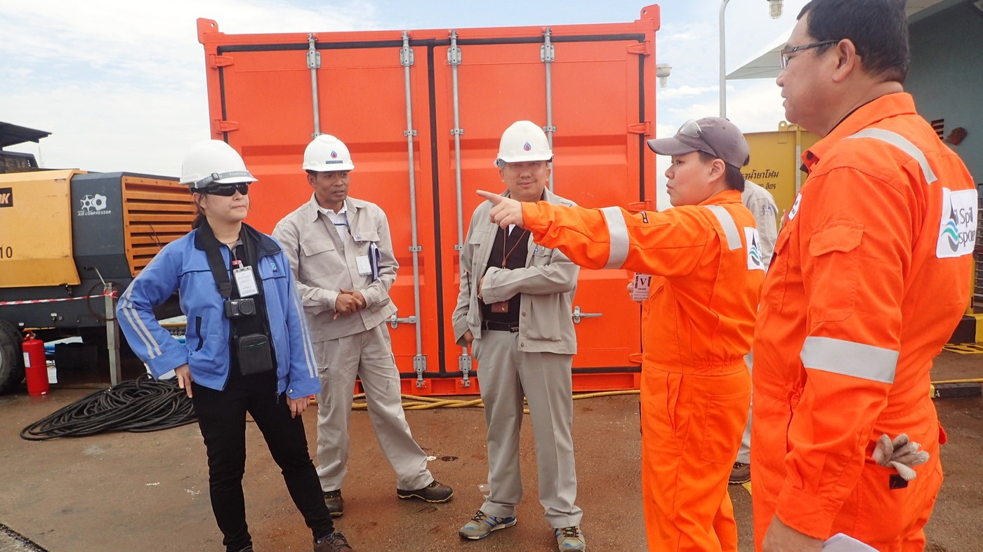 An oil spill response instructor points out directions during a training session, with participants in orange safety gear standing next to emergency response equipment.