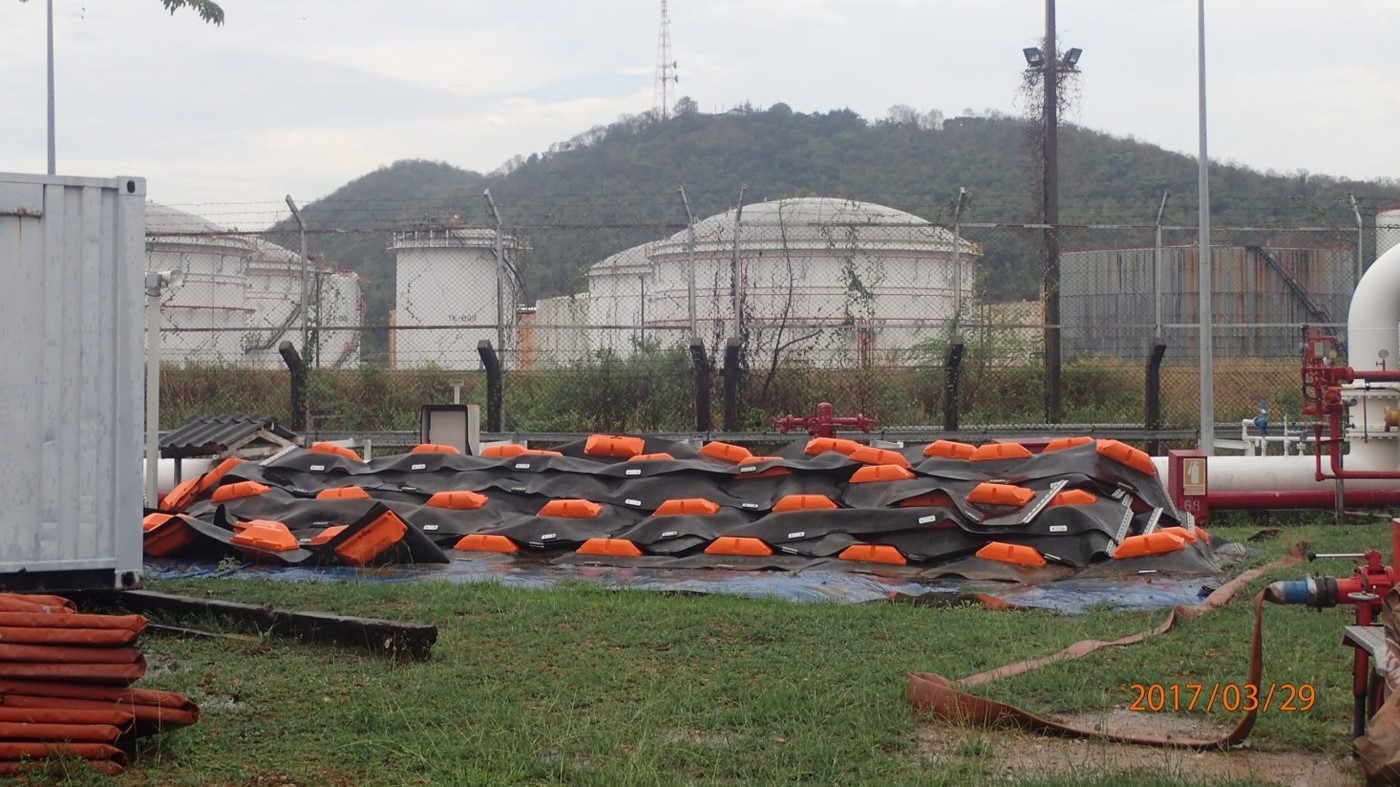 A collection of oil spill containment booms is laid out on a tarpaulin near large white oil storage tanks, with a backdrop of green hills and cloudy skies.
