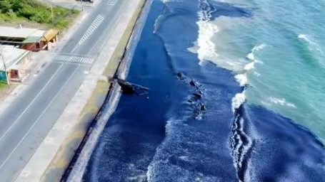 An aerial photograph of a coastline in Tobago, showing crude oil washing onto the shore and roads, demonstrating the scale of contamination and the need for rapid response.