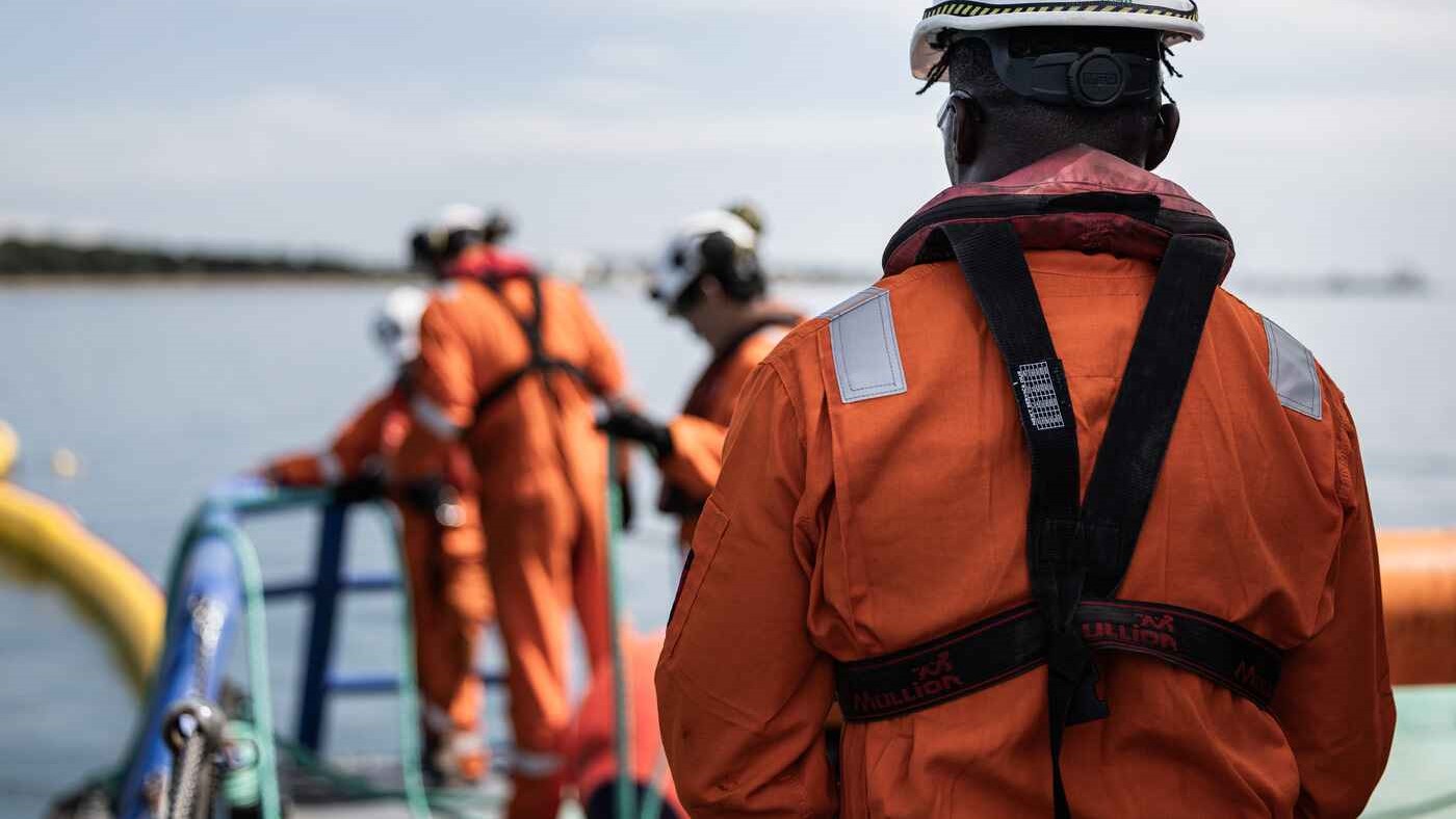 Response team members deploy orange containment boom from a dock as part of a harbour-based training scenario—demonstrating equipment readiness and response coordination.