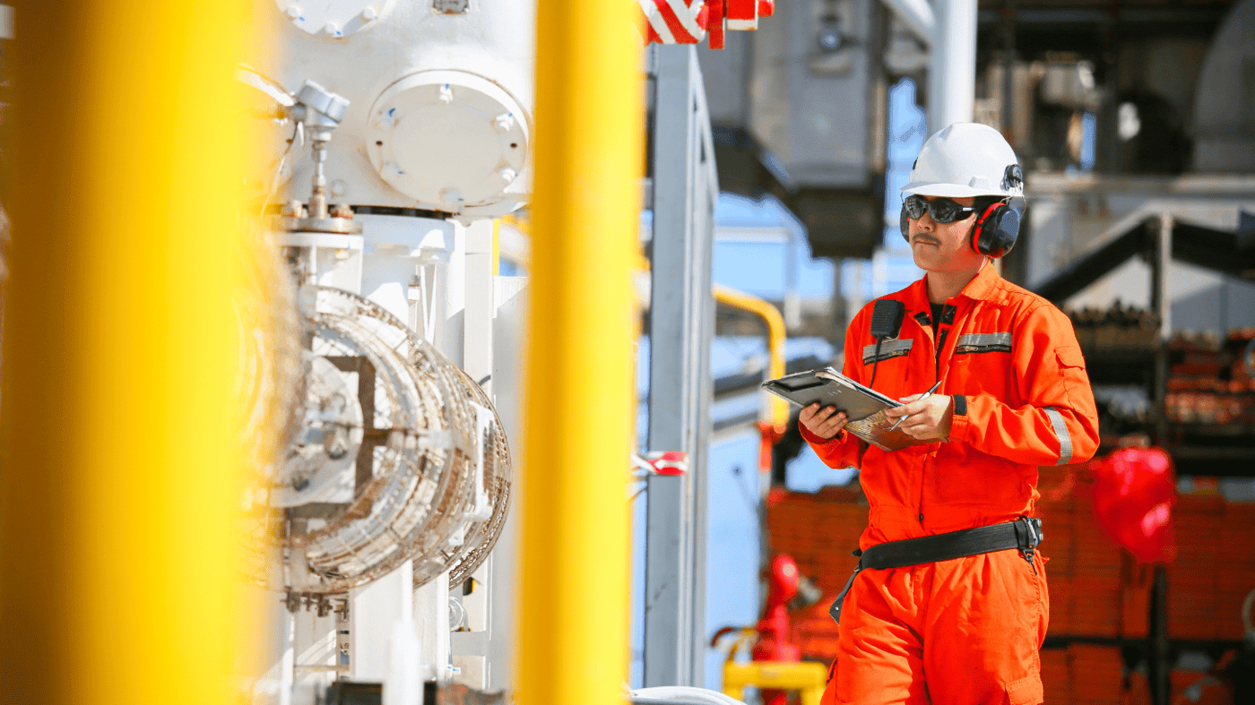 A safety officer in orange PPE conducts an inspection with a digital tablet on an offshore platform. The image illustrates the proactive assessment and operational oversight involved in effective oil spill contingency planning, where routine inspections play a key role in identifying risks and readiness gaps.