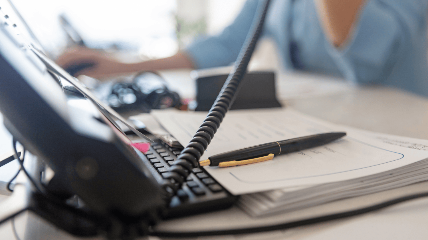 A focused view of a telephone with a coiled cord, a black and gold pen, and a notepad with notes, symbolising key tools for crisis communication. In the background, a blurred professional is engaged in a phone call, emphasising the importance of timely and strategic communication during a crisis.