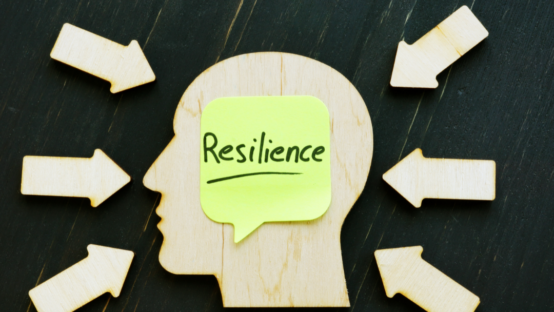 A symbolic visual of mental strength and adaptability, with a human profile cut-out and the word “Resilience” highlighted on a sticky note. Arrows pointing inward depict external pressures and decision-making stress, often experienced during crisis scenarios. This image reflects the need for crisis leaders to build mental resilience beyond virtual simulations.