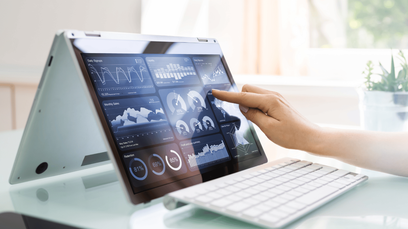 A convertible laptop with a touch screen shows various data analytics, including charts, graphs, and performance indicators. A hand is seen pointing at the screen, highlighting interactive decision-making and digital monitoring. The workspace includes a keyboard and a plant in the background.