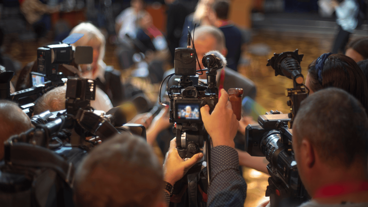 A chaotic media scene with numerous journalists and camera operators capturing a press conference. Professional video cameras, microphones, and reporters with recording equipment surround a speaker, whose blurred figure is visible in the centre. The atmosphere suggests an important announcement or crisis response briefing.