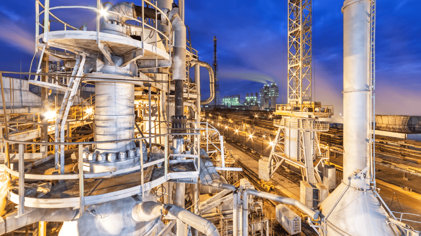 sprawling petrochemical plant glowing under artificial lights against a deep blue evening sky. The facility includes industrial towers, extensive piping systems, and complex processing units, likely involved in ammonia or chemical production. Smoke or vapour can be seen rising from some sections, indicating active operations.