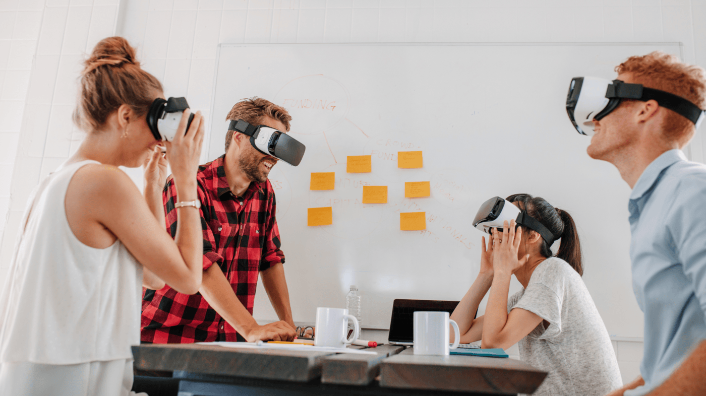 Four individuals in a modern office setting participate in a virtual reality training session, wearing VR headsets. They are seated around a table with laptops, coffee mugs, and notes, suggesting a collaborative learning environment. A whiteboard in the background contains handwritten notes and sticky notes, indicating brainstorming or training exercises.