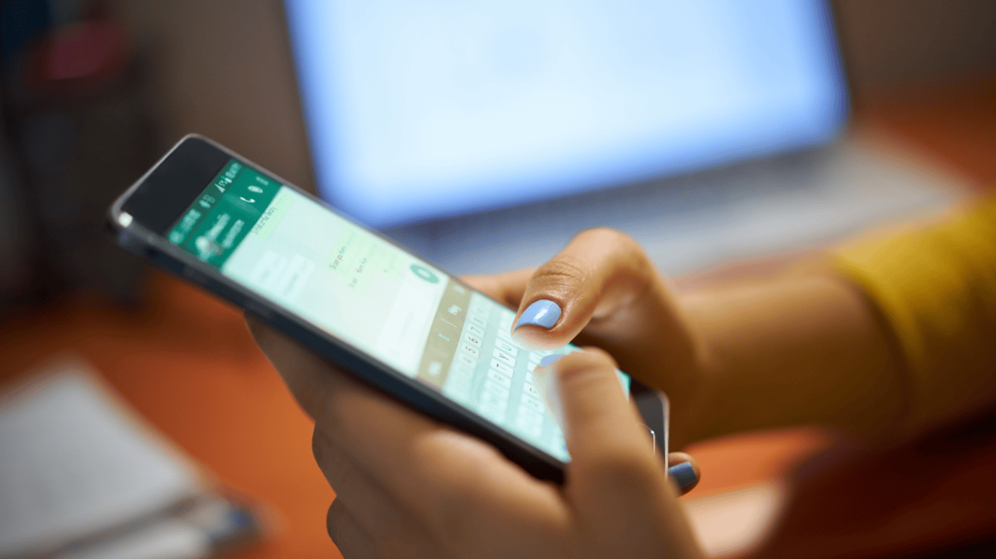 A close-up shot of a person's hands holding a smartphone, typing a message on WhatsApp. The background features a blurred laptop screen, indicating multitasking or remote communication. The image conveys modern digital communication and connectivity.
