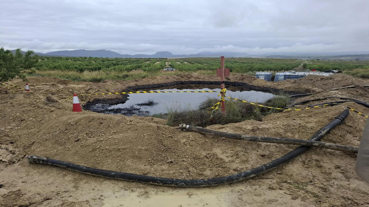 A recovery pit filled with oil sits in an agricultural field, encircled by hazard tape and traffic cones. Black hoses run across the ground, with IBC tanks stacked in the background under a cloudy sky.