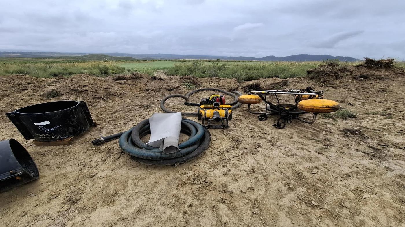 A skimmer unit, hose, portable pump, and connector parts are arranged on a dirt surface near grassy terrain. The equipment is ready for use in oil recovery operations in a remote inland setting.