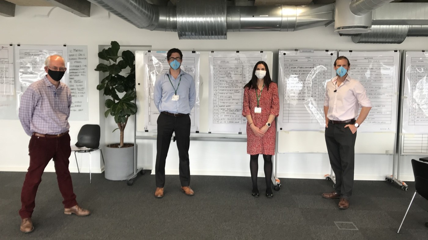 A crisis management training session at Total EP Denmark, featuring a small team of professionals standing in front of incident response boards. Participants are wearing masks, following COVID-19 protocols, and discussing emergency procedures.