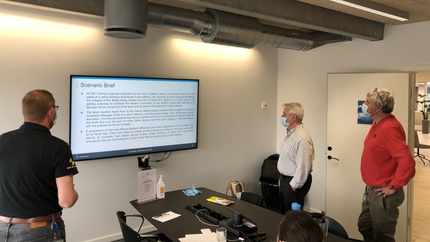 A crisis scenario briefing session at Total EP Denmark, where professionals are analysing a scenario displayed on a large screen. The briefing outlines emergency procedures and response actions in a crisis situation.