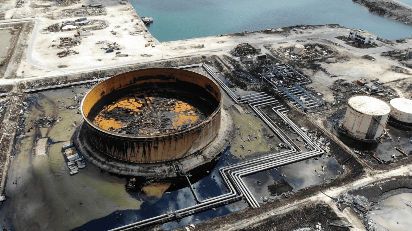 Aerial photograph of an oil storage facility devastated by Hurricane Dorian in the Bahamas. A large, rusted oil tank with a collapsed roof is surrounded by contaminated water, pipelines, and debris. Other storage tanks show signs of severe structural damage, while the surrounding area appears flooded and littered with wreckage.