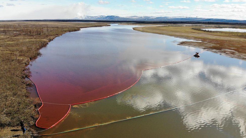 A river affected by an oil spill, with a large reddish slick spreading across the water. Red containment booms are deployed along the shoreline and across the river to prevent further contamination. A small boat is assisting in the cleanup. The surrounding landscape includes barren land and distant mountains under a partly cloudy sky.