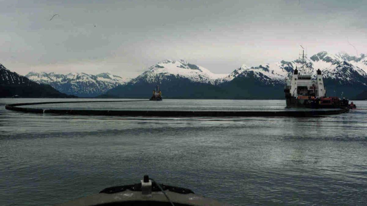 Two vessels encircling oil with a large containment boom against the backdrop of snowy mountains. This large-scale containment strategy was used to prevent oil from spreading further in Prince William Sound.