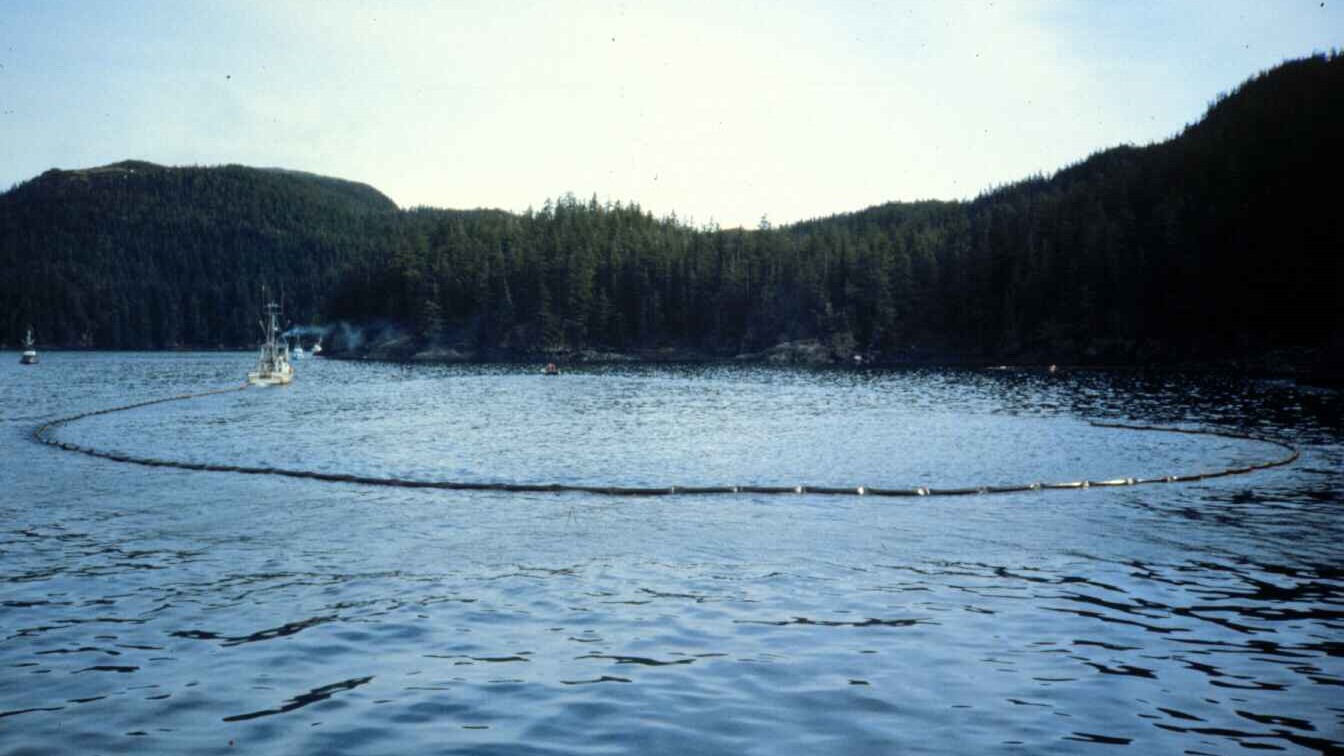  A vessel working to deploy an oil containment boom in calm waters with forested shoreline in the background. This containment strategy was crucial in limiting the spread of crude oil following the Exxon Valdez spill.