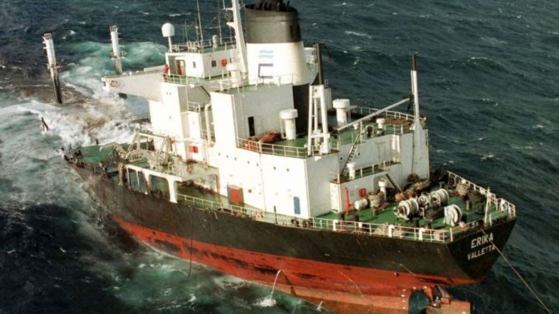 The MV Erika, a heavily damaged oil tanker, is seen partially submerged in turbulent waters. The vessel’s stern is sinking while the bow remains afloat, highlighting the severity of the maritime disaster. The ship's name, “Erika Valletta,” is visible on the hull, marking the moment before it fully broke apart and spilled thousands of tonnes of oil off the coast of France.